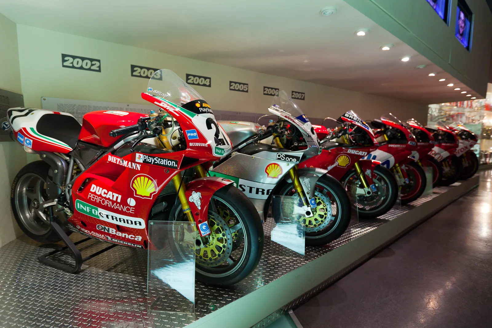 A row of red and white Ducati racing motorcycles on display in the Ducati Museum at the Borgo Panigale factory in Bologna, each bike under a sign giving its year