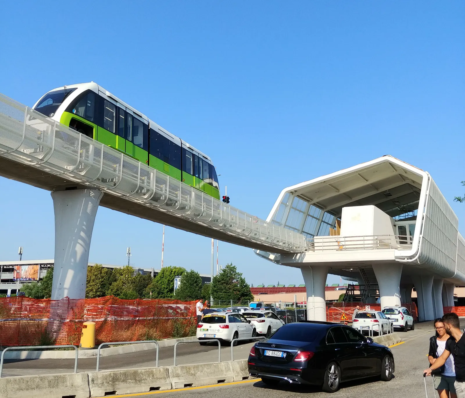 A green and white Marconi Express monorail car running along its raised concrete track into the elevated airport station at Bologna, cars parked below