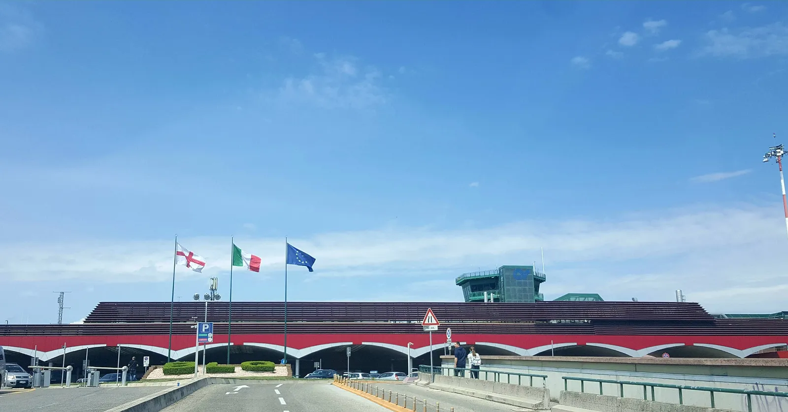 The front of the passenger terminal at Bologna Guglielmo Marconi Airport, a long low building with a red arched canopy along its frontage, flags flying out front and the control tower behind