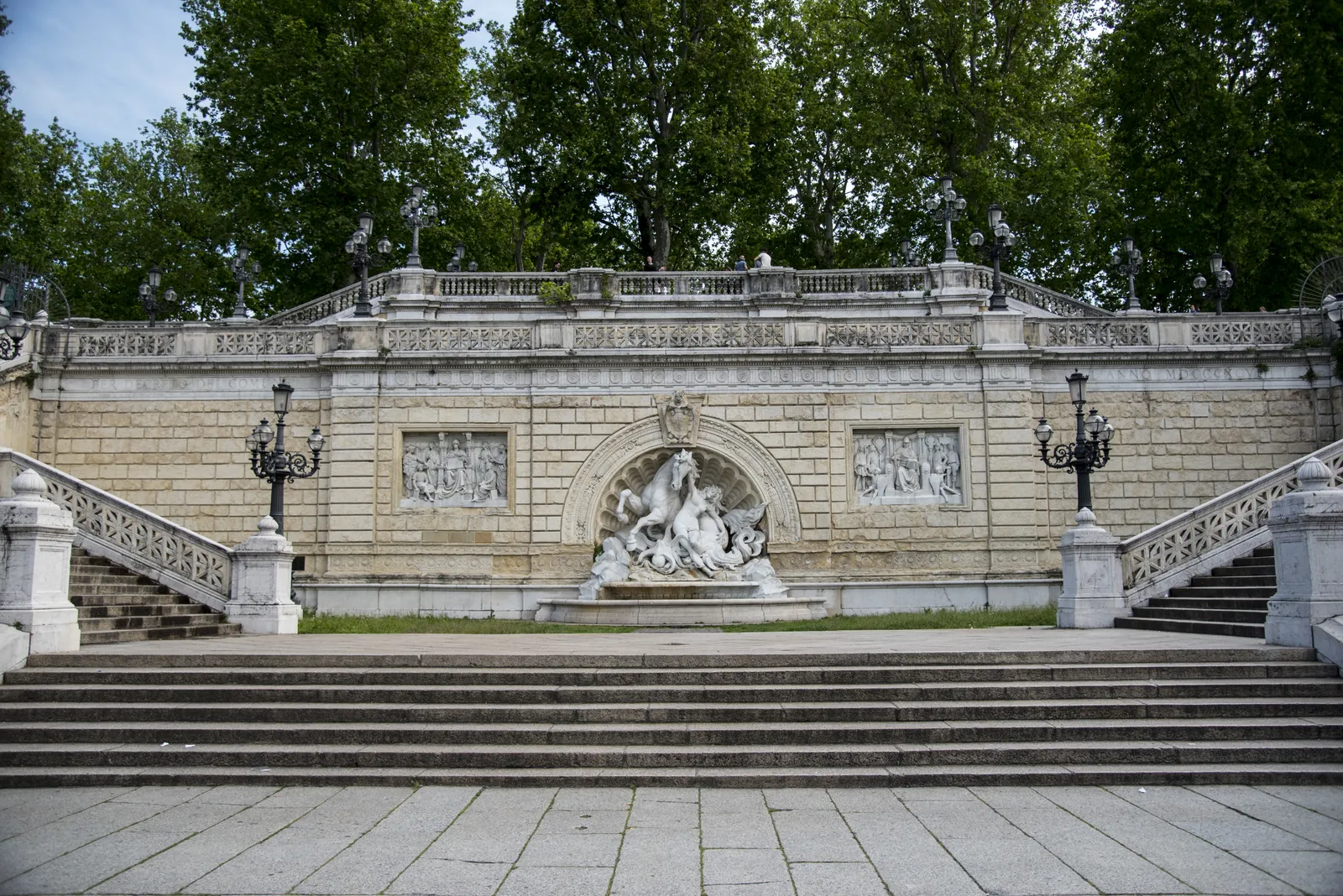 The Pincio staircase in Bologna, twin flights of stone steps rising either side of a sculpted fountain group and carved relief panels, with the trees of the Montagnola park above