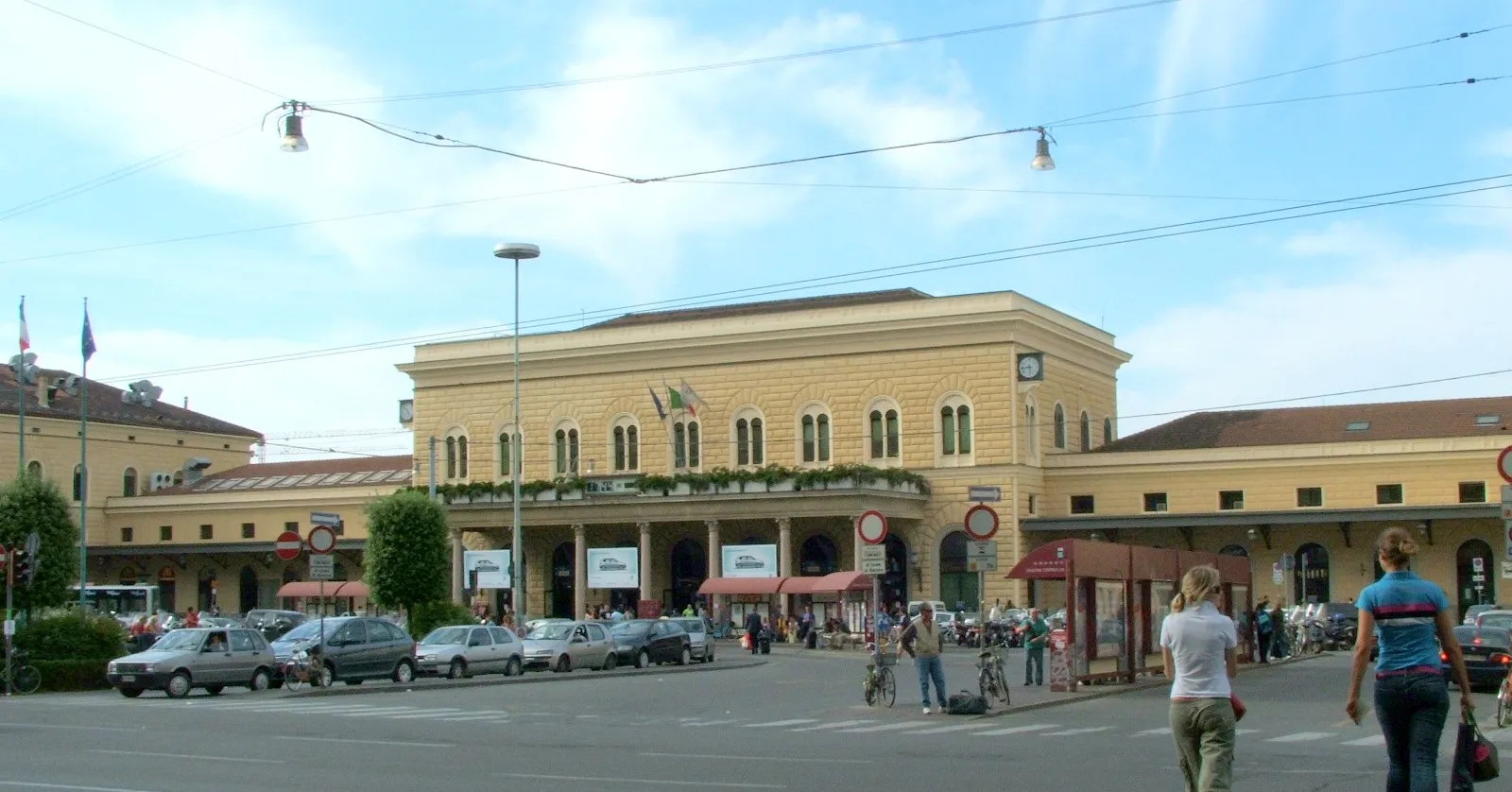 The pale yellow front of Bologna Centrale railway station across Piazza delle Medaglie d'Oro, with arched windows, a clock on the right of the upper storey and a colonnaded entrance porch