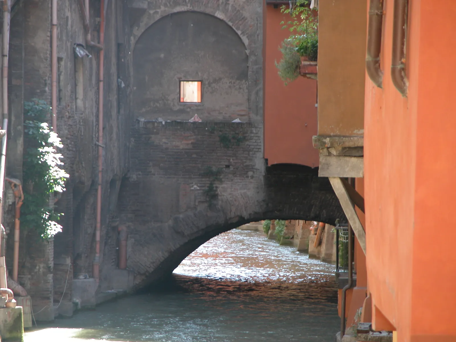 A canal in central Bologna running under an arch between old houses, with the small square window of Via Piella set in the brick wall above the water