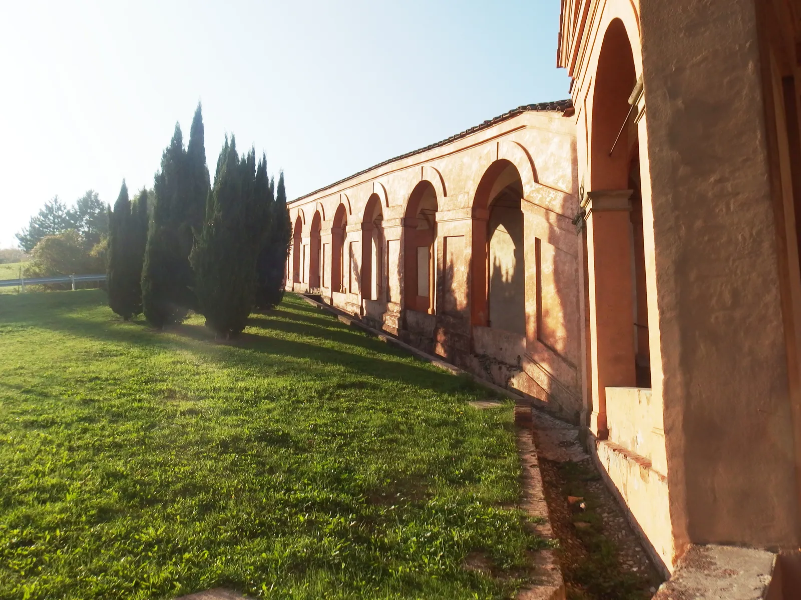 A long row of brick arches of the San Luca portico running along a grassy hillside outside Bologna, lined by tall cypress trees in low sunlight
