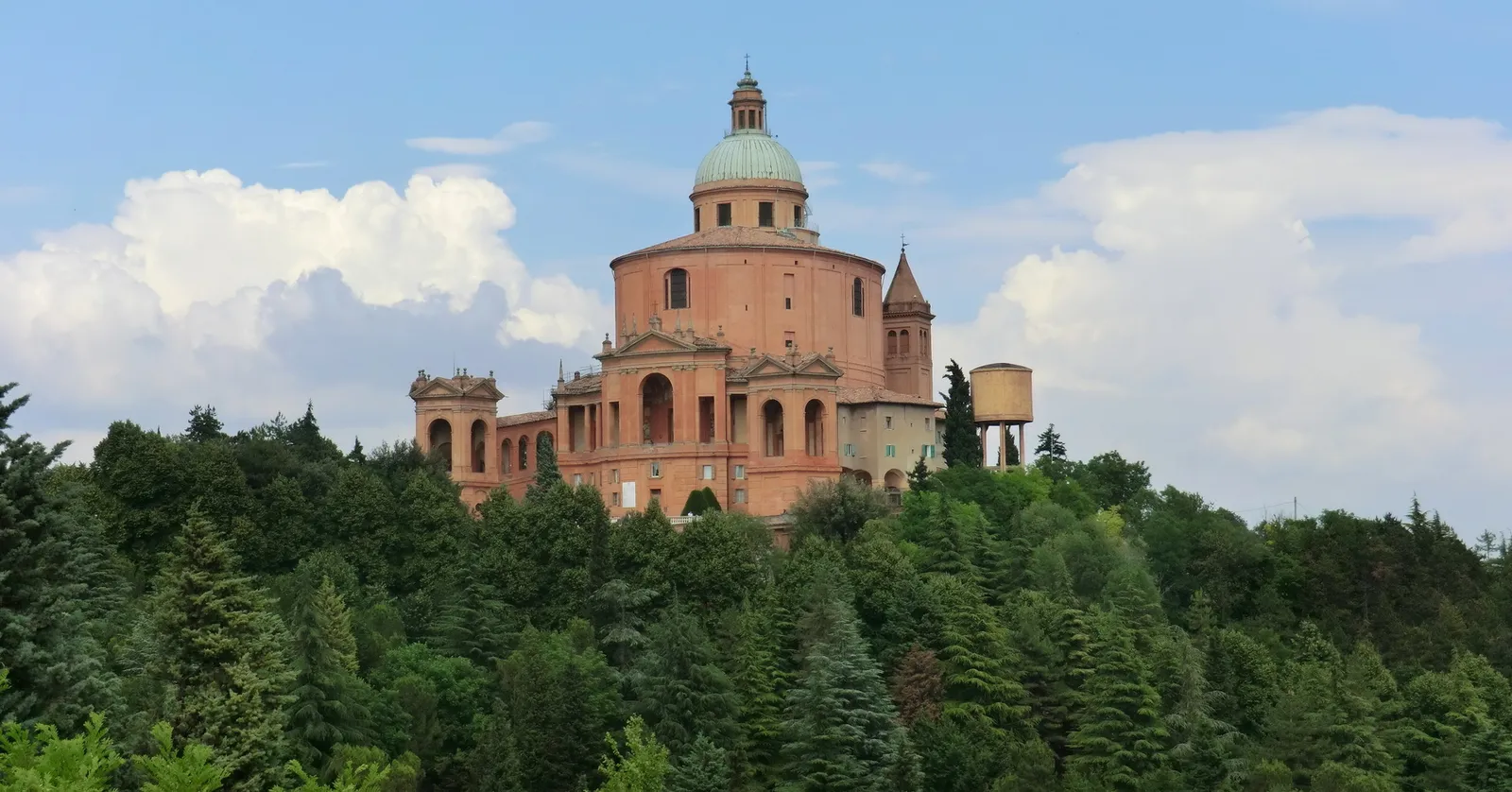 The Sanctuary of the Madonna di San Luca above Bologna, a domed pink-brick church with a green copper cupola rising over a wooded hilltop under a blue sky with white clouds