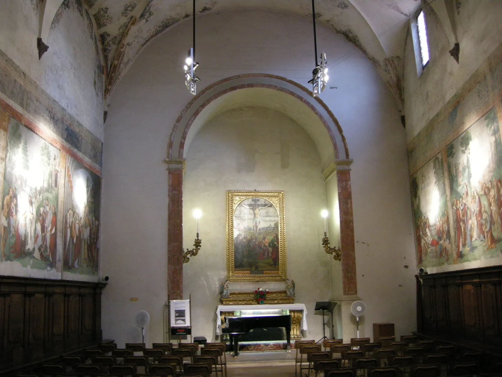 The interior of the Oratory of Santa Cecilia on Via Zamboni in Bologna, its side walls covered in Renaissance fresco panels, rows of chairs and a piano in front of the altar
