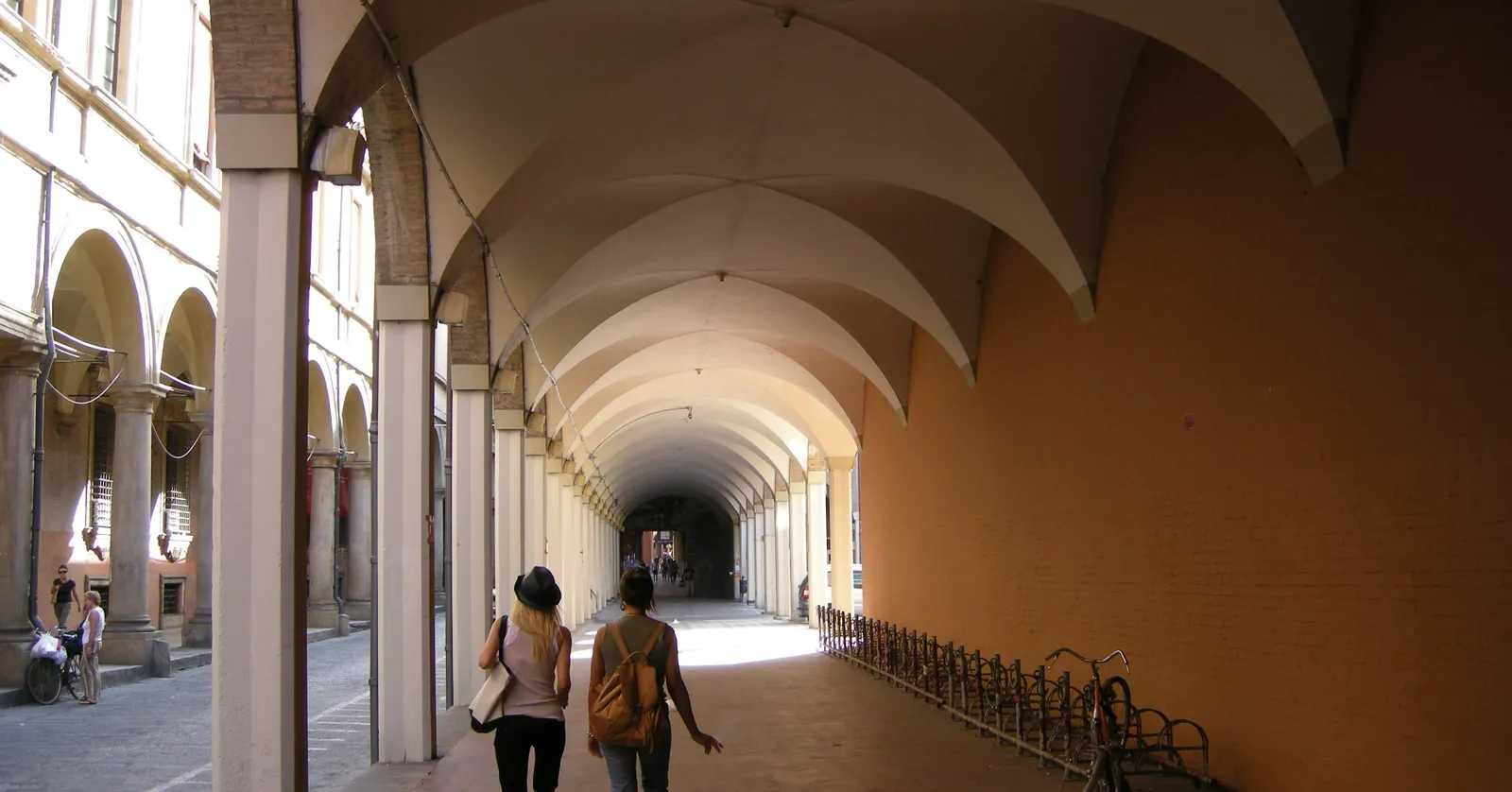 Two people walking under the long vaulted arcade of Via Zamboni in Bologna's university quarter, a row of white columns on one side and an orange wall with bicycle racks on the other