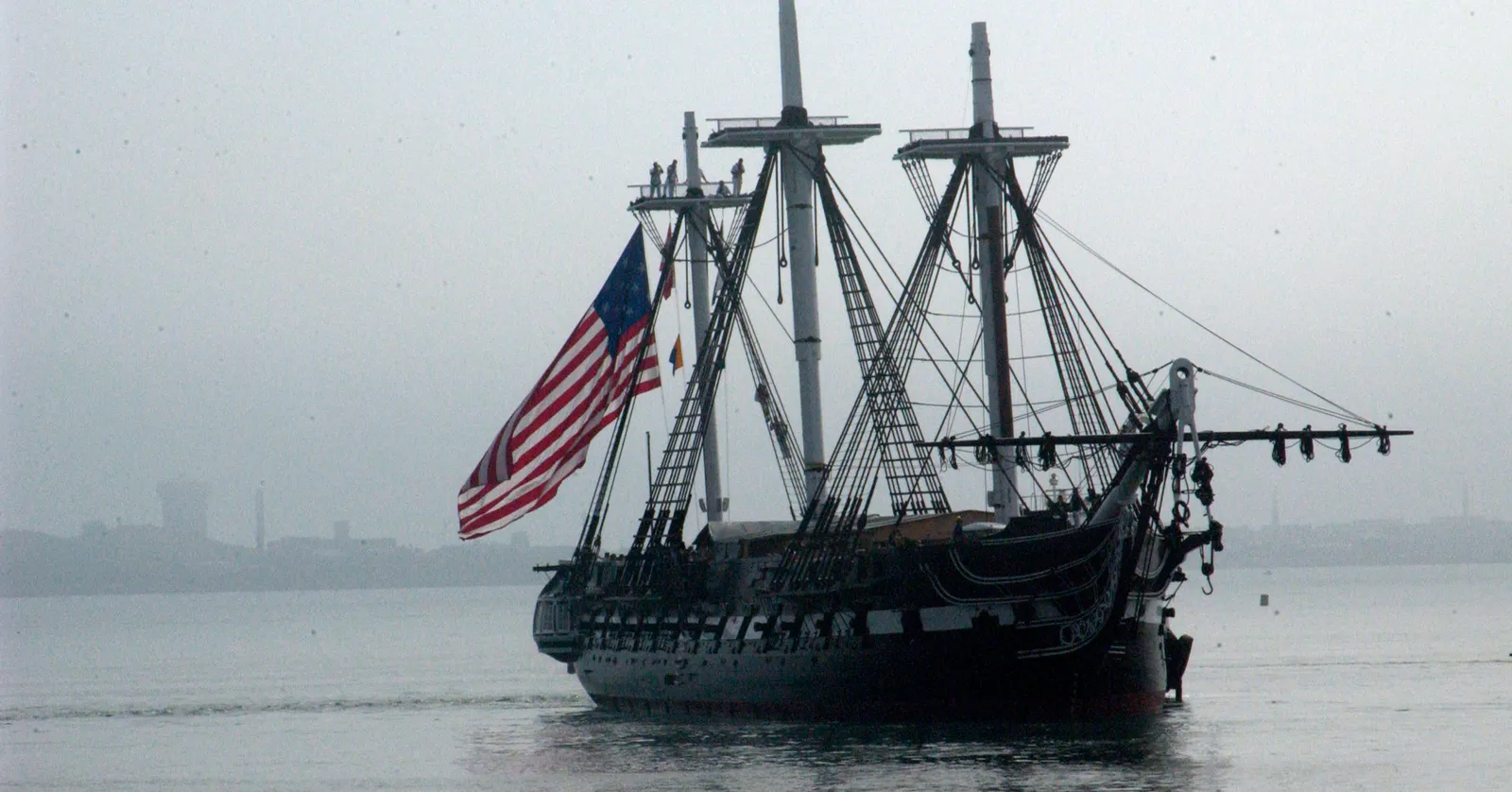 USS Constitution on the water in Boston Harbor, a three-masted wooden warship with a black hull, white gun stripe and a large United States flag flying from her rigging