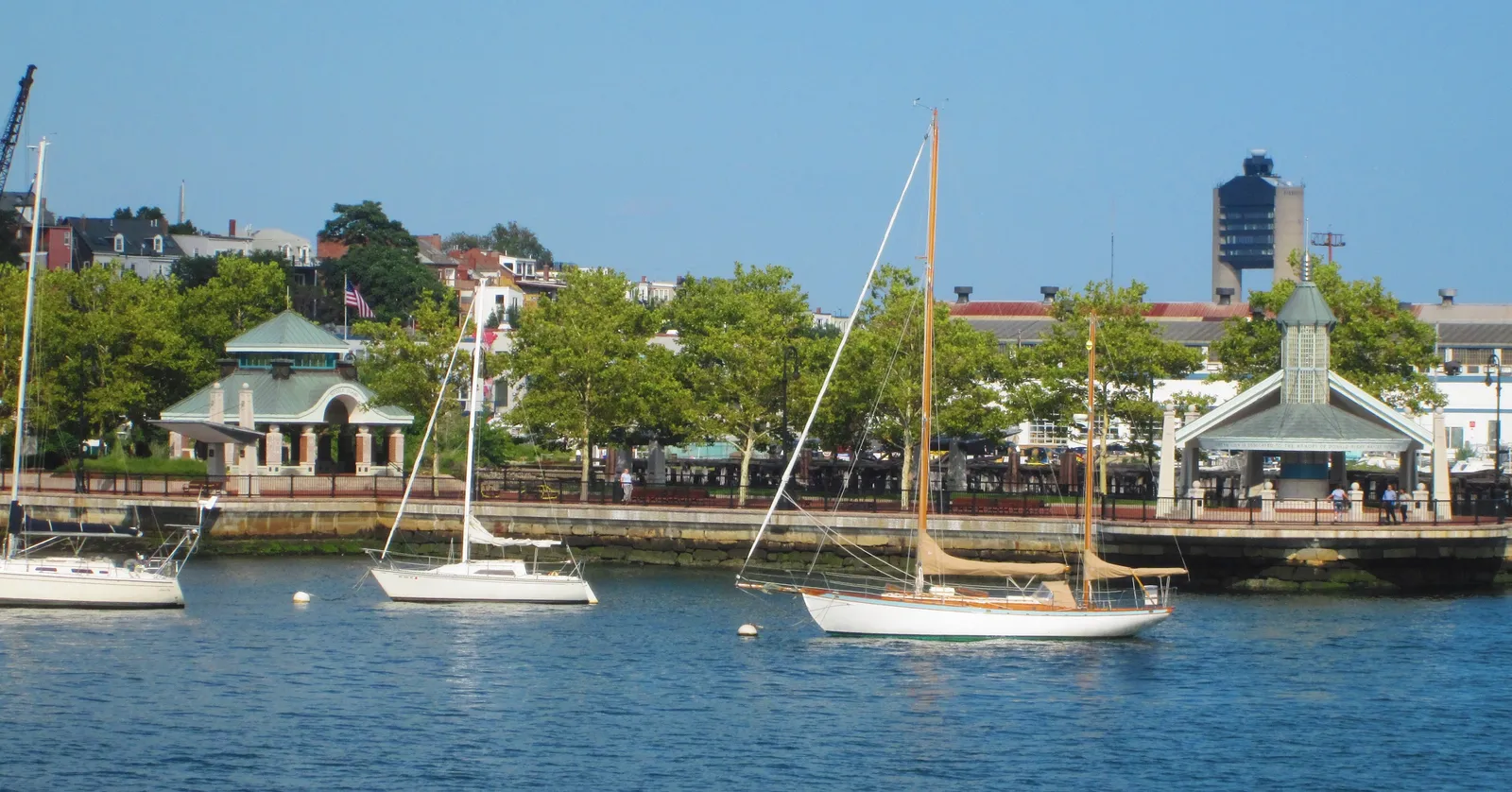 Sailboats moored off Piers Park in East Boston, with the park's pavilions on the waterfront and Boston Logan airport's control tower rising behind the trees