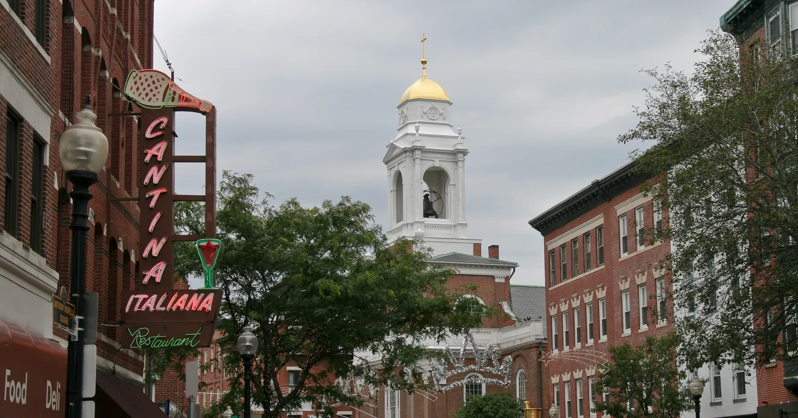 Hanover Street in Boston's North End, a neon Italian restaurant sign hanging over the pavement and a white church tower with a gilded dome standing further up the street between brick blocks