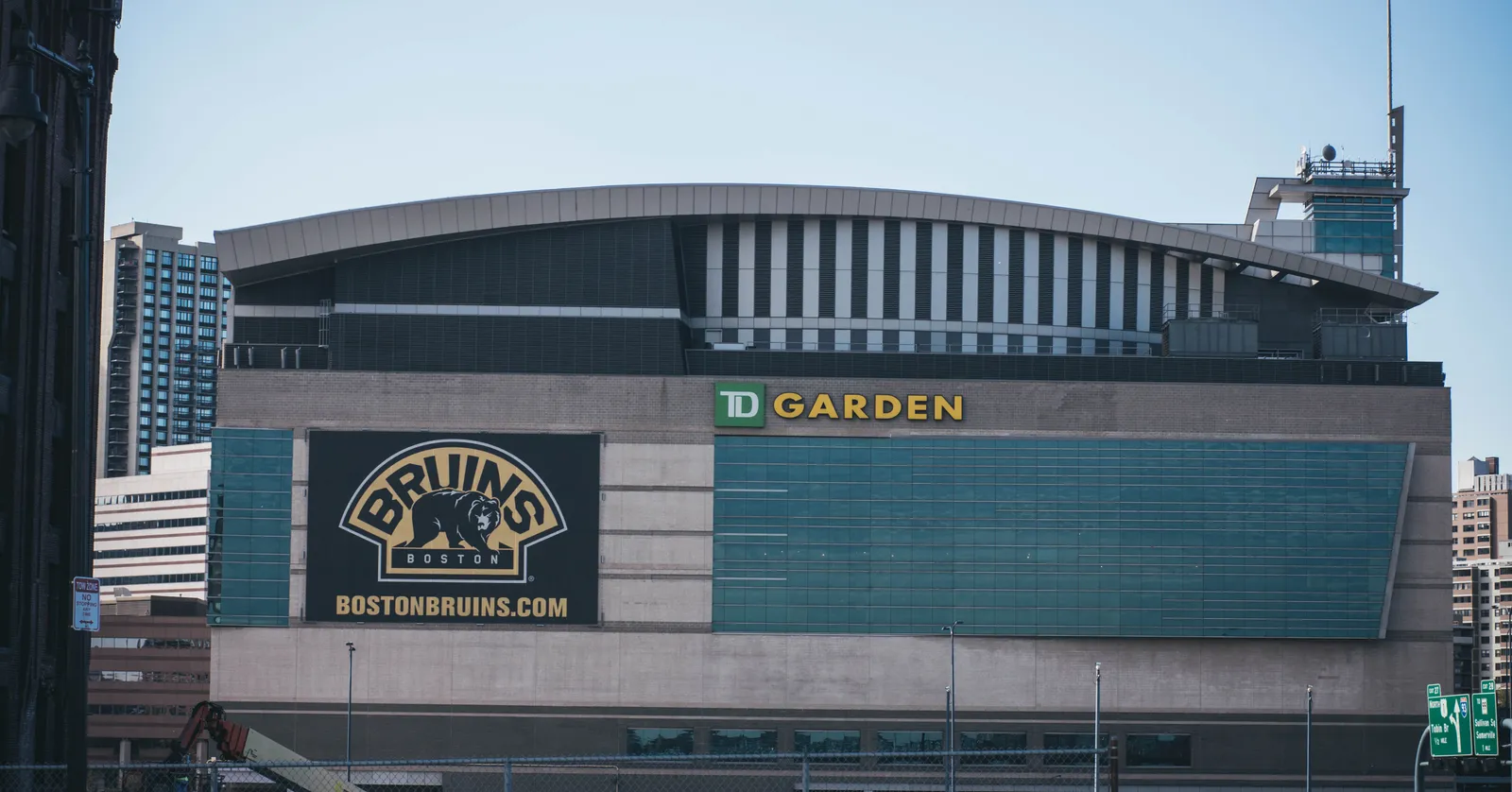 The curved roof and green glass front of TD Garden in Boston, with the arena's name across the parapet and a Bruins crest on the panel below it