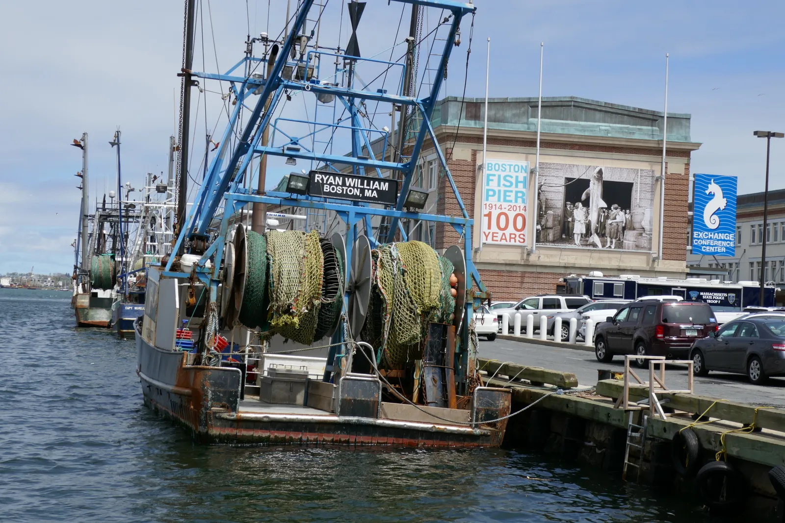 A working trawler tied up at the Boston Fish Pier with its nets and winch drums on deck, and a brick pier building behind carrying a Boston Fish Pier banner