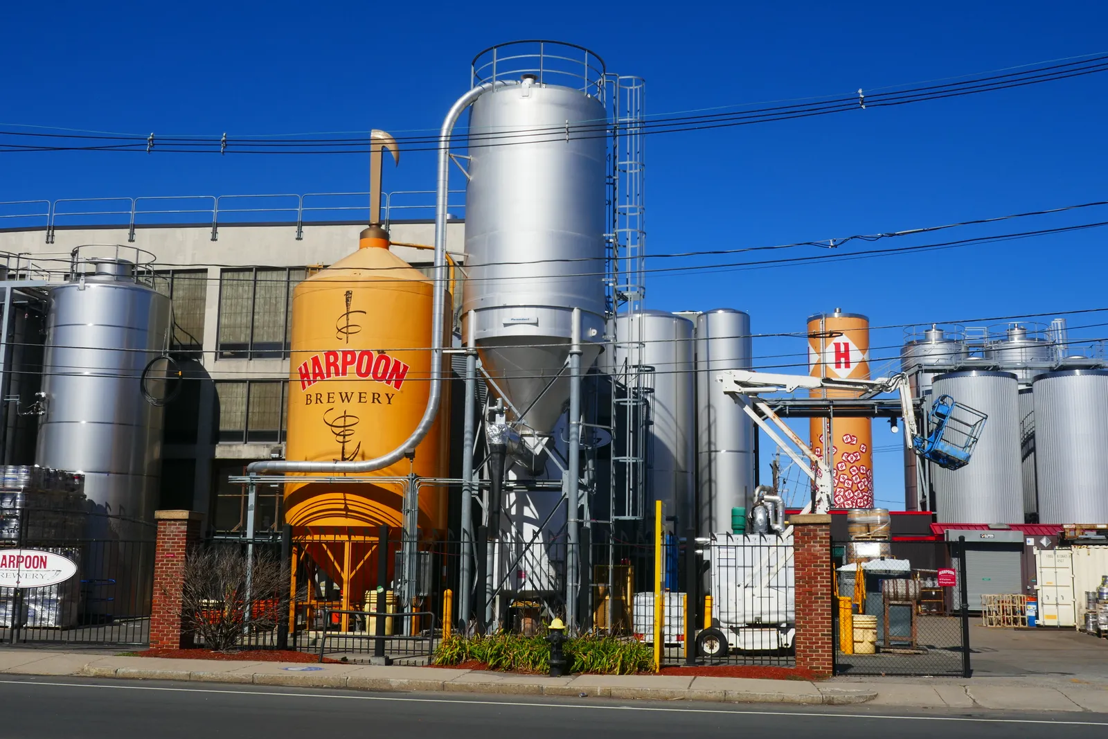 The Harpoon Brewery on Northern Avenue in Boston, steel fermentation tanks and a painted kettle standing behind railings at the pavement edge under a clear sky