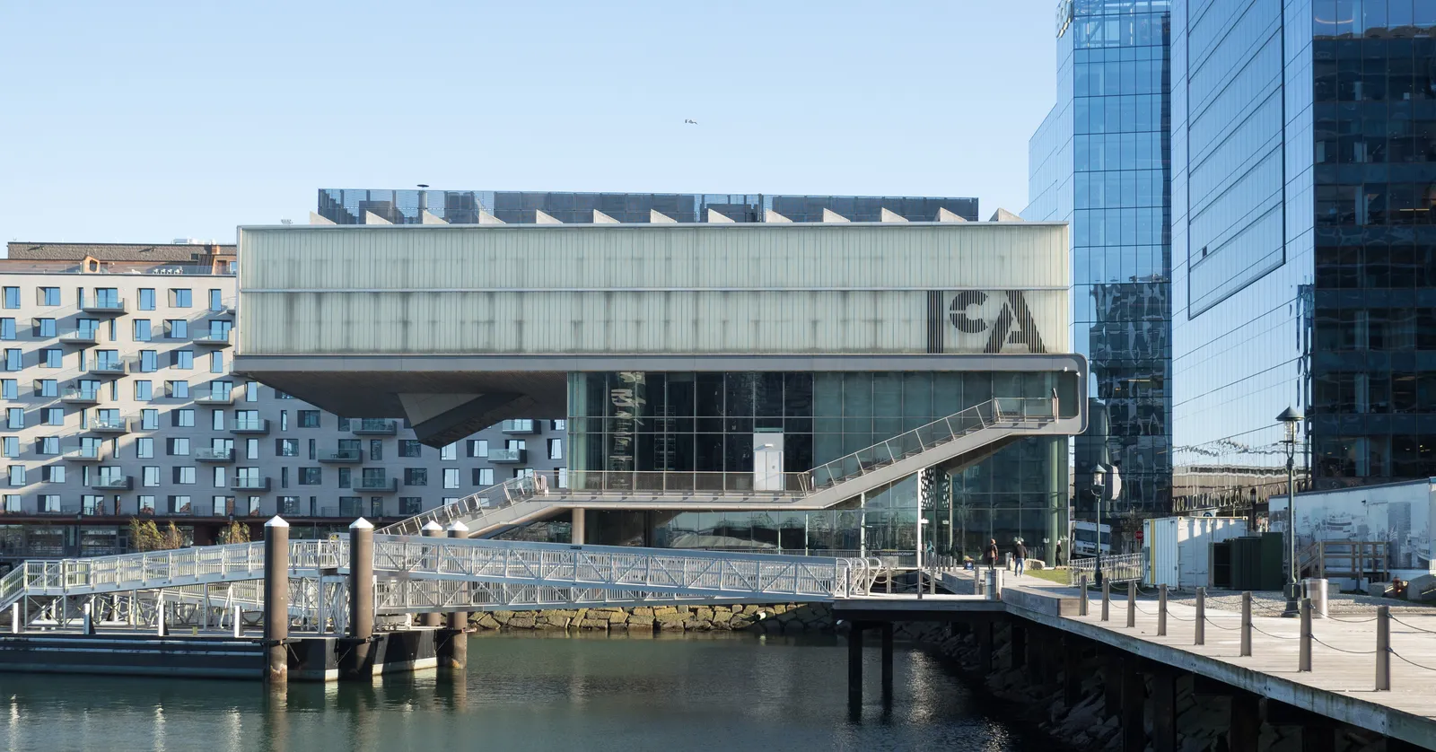 The Institute of Contemporary Art in Boston seen across the water, its upper gallery projecting out over the waterfront path with the building's initials on the glass front and a stepped grandstand running down to the harbour