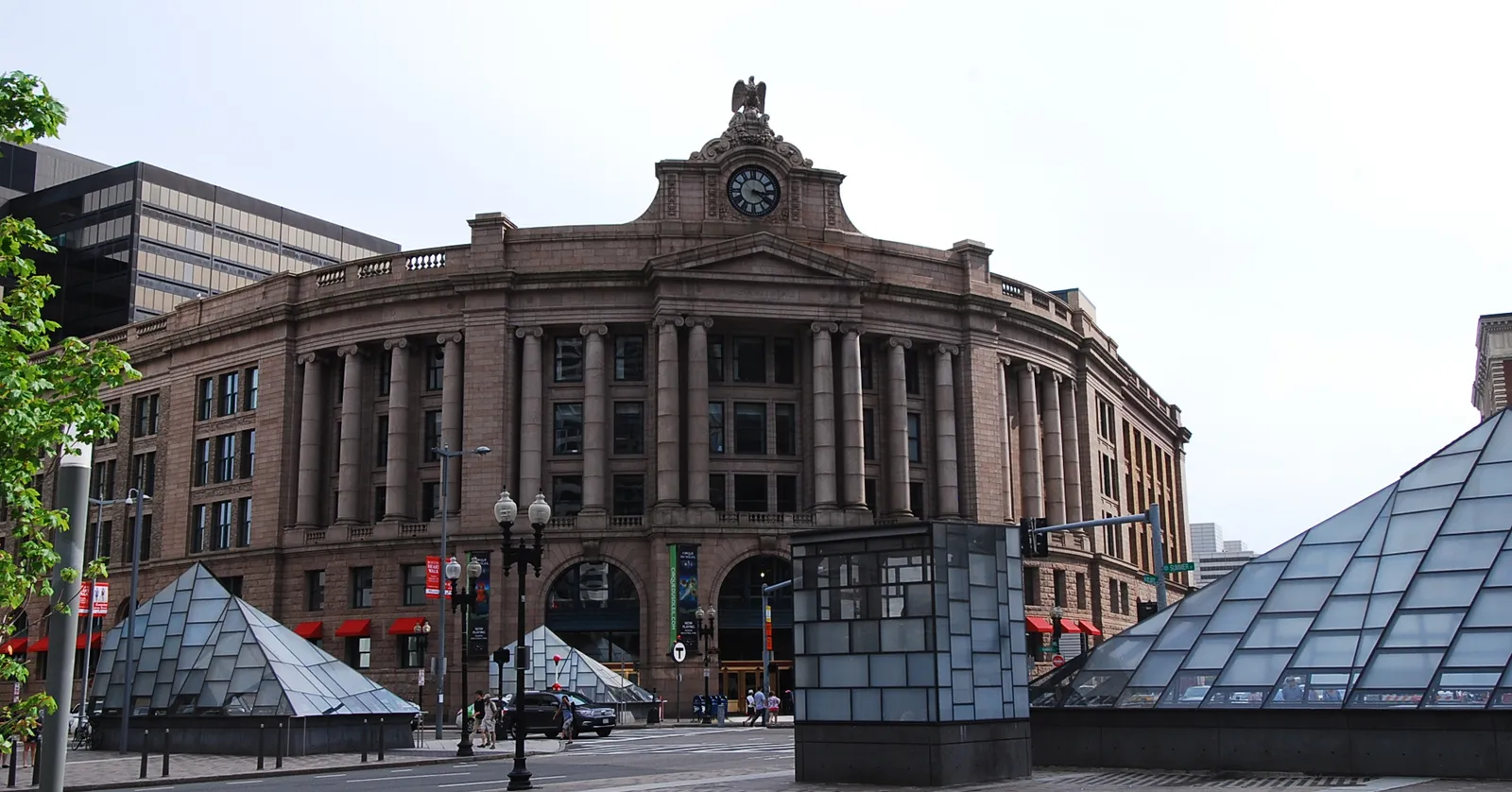 The curved granite front of Boston's South Station head house on Dewey Square, a clock and a stone eagle above the central bay and glass pyramid entrances standing on the plaza in front