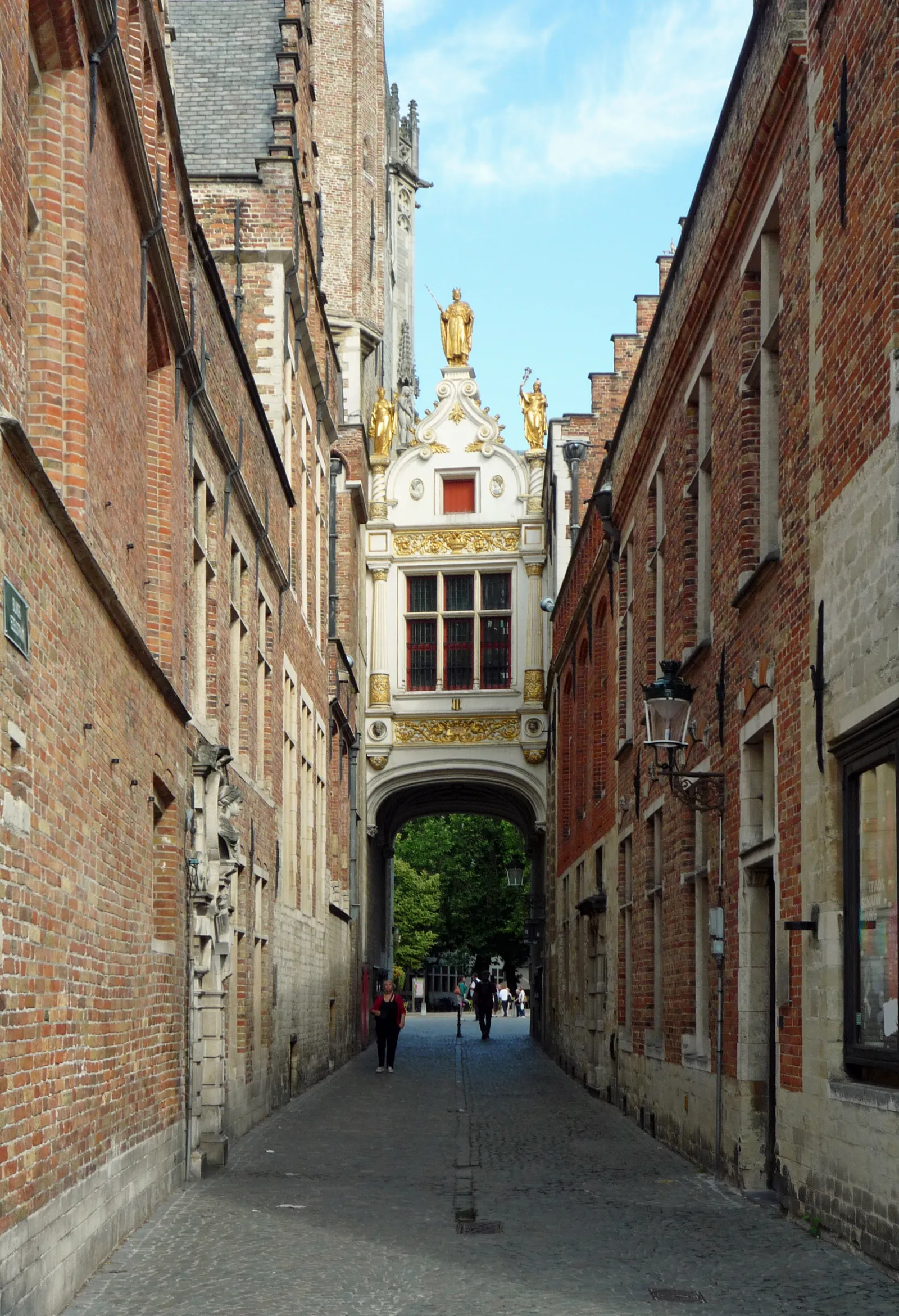 A narrow brick lane in Bruges running under a white stone arch topped with gilded statues, cobbles underfoot and people walking through towards daylight at the far end