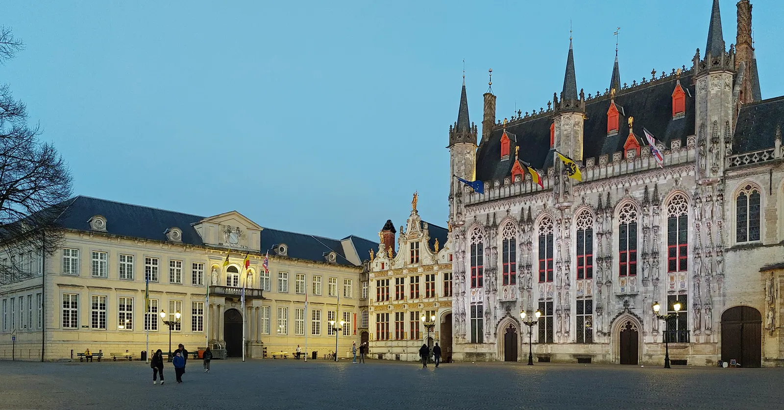 Burg square in Bruges at dusk, the floodlit Gothic city hall with its turrets and statue niches on the right and the long classical front of the provost's house on the left, the paving empty in front of them