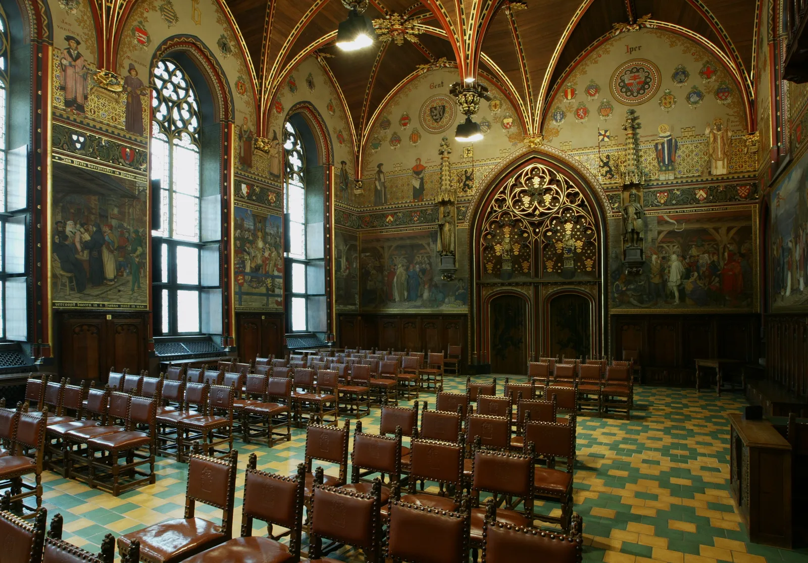 The Gothic Hall inside Bruges city hall, a ribbed timber vault painted in gold and red over walls of large historical murals, with rows of leather chairs on a green and yellow tiled floor