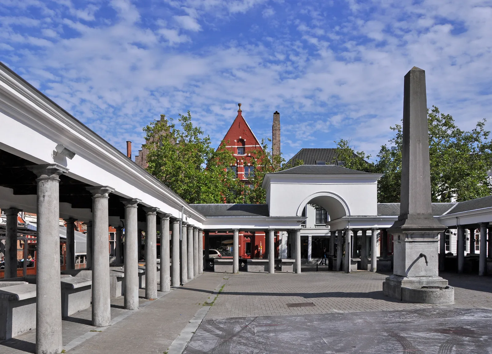 The fish market colonnade in Bruges, two rows of slim stone columns carrying a low roof over long stone selling slabs, with a stone obelisk standing in the open paving between them