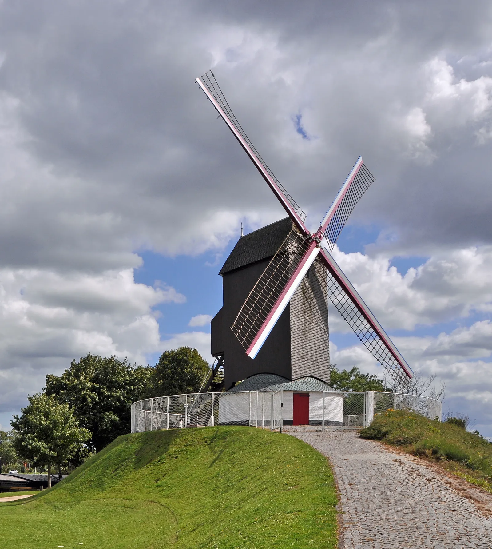 A black timber windmill in Bruges on a grassy earth mound with a cobbled path curving up to it, its four sails set against a sky of heavy cloud