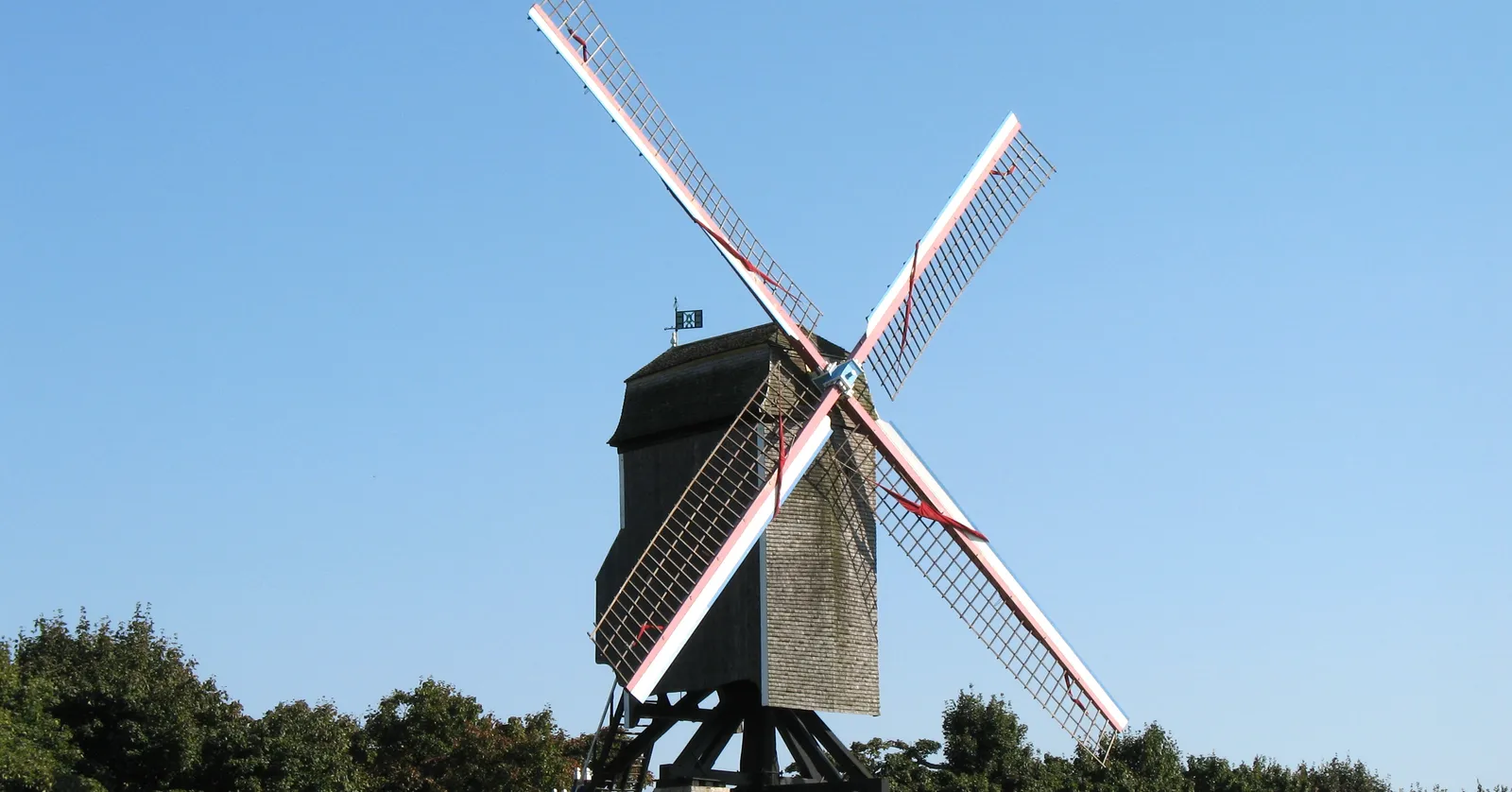 A wooden post windmill in Bruges standing on its brick base against a clear blue sky, four lattice sails turned to the wind above a line of trees