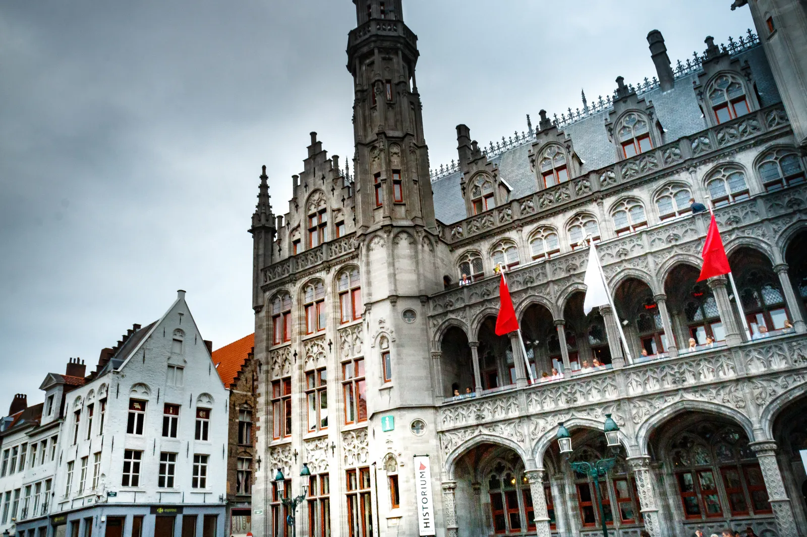The Historium building on the Markt in Bruges, a tall neo-Gothic front of pale stone and red brick with a slender turret, standing beside the arcaded provincial government building