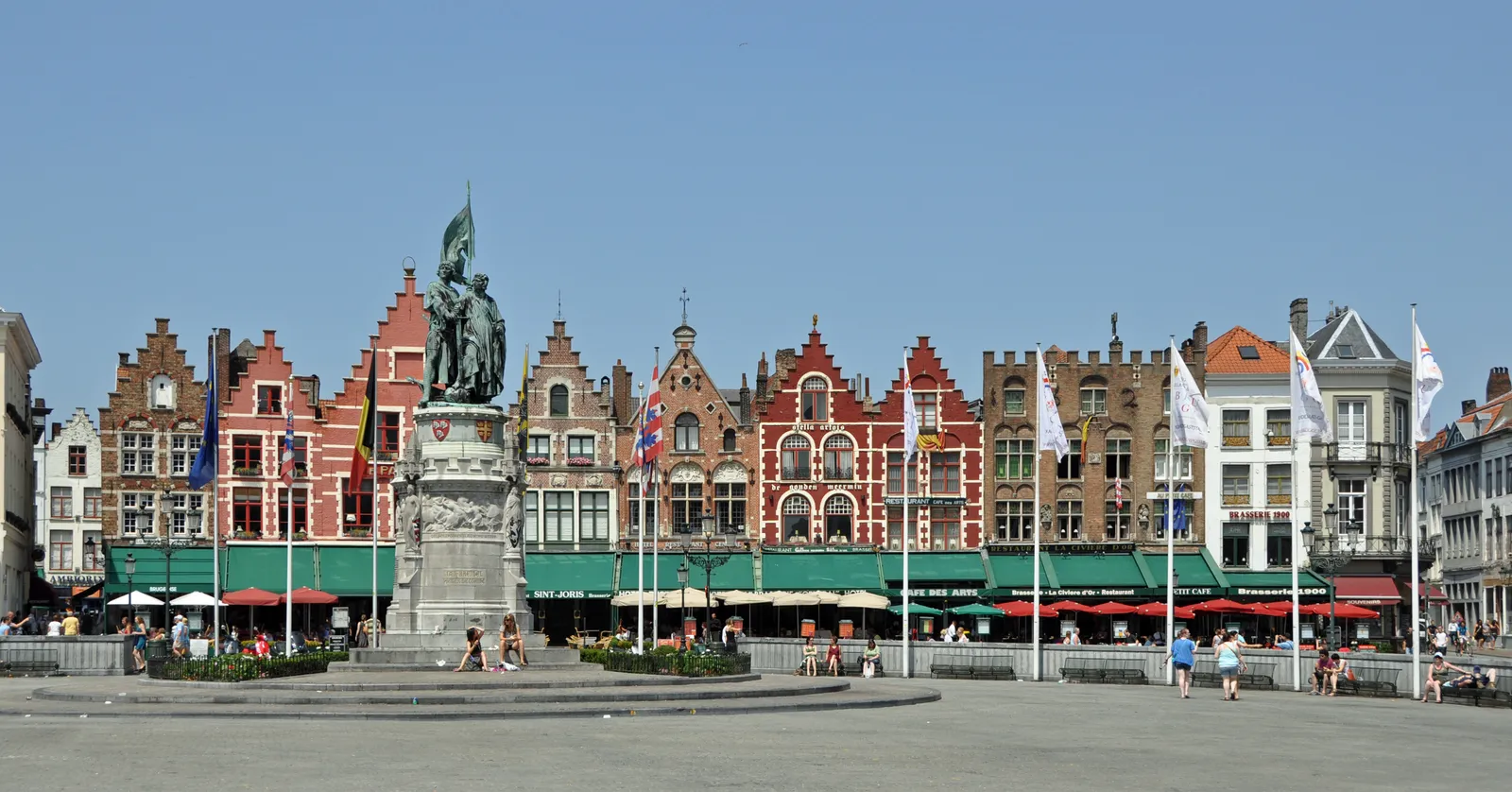 The north side of the Markt in Bruges, a long row of stepped-gable houses painted in reds, greens and ochres above green cafe awnings, with a bronze statue on a stone plinth in front of them