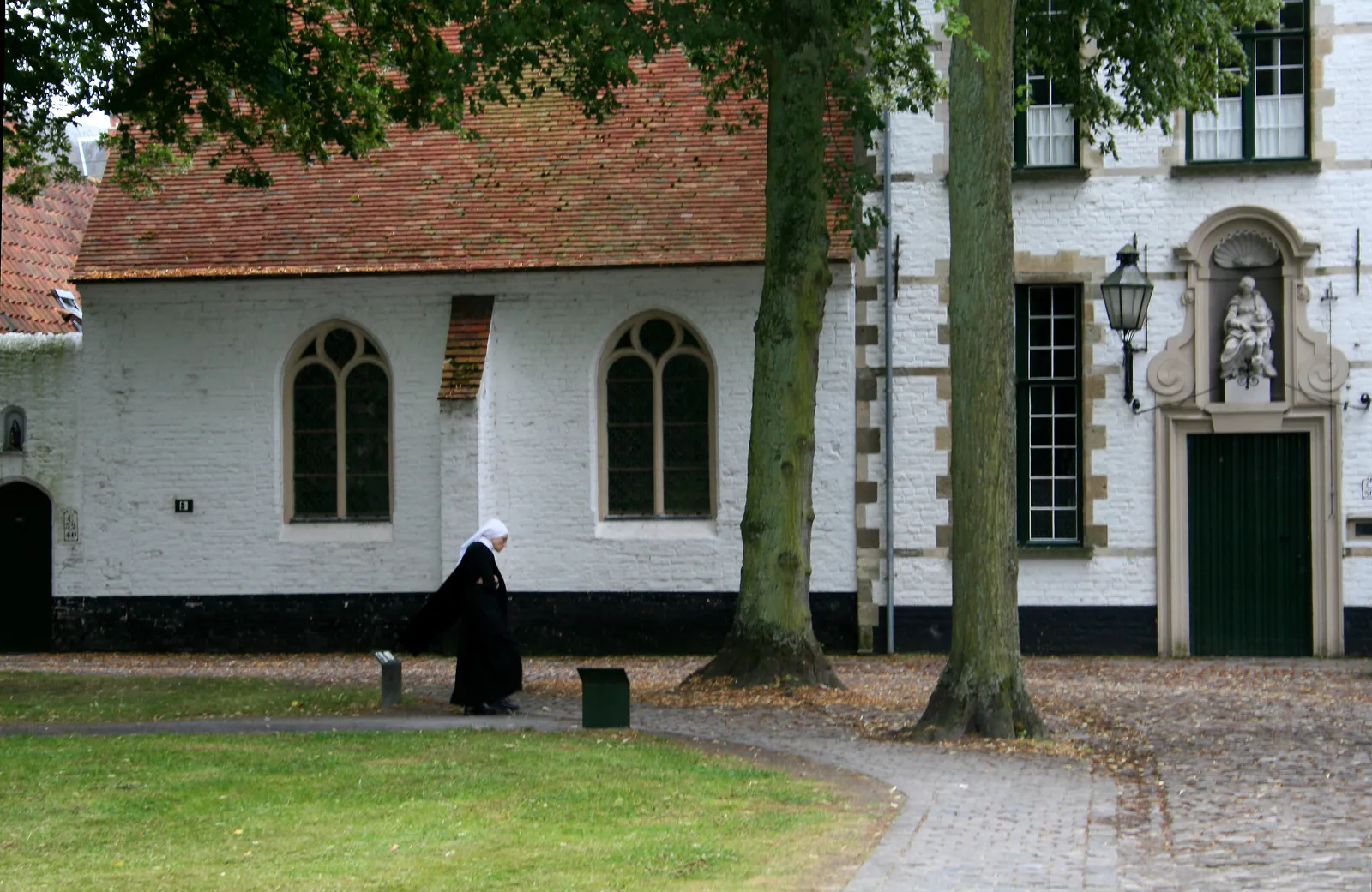 A sister in a black habit and white veil walking past the whitewashed chapel and gabled houses of the Beguinage in Bruges, with a gravel path and lawn in front of them