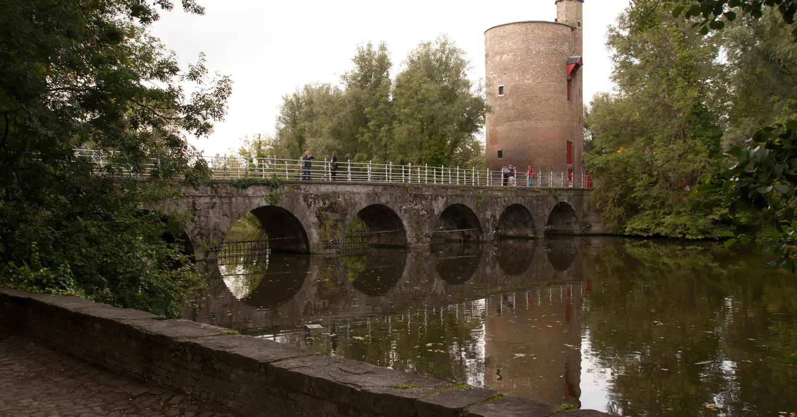 The arched stone bridge over the Minnewater in Bruges reflected in still water, with a squat round brick tower standing among trees on the far bank