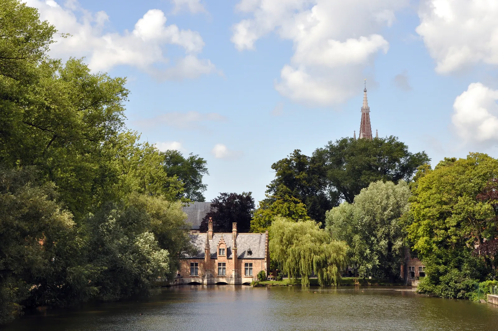 The Minnewater in Bruges, a still rectangular lake edged with willows, a small brick gabled lodge at the water's edge and a church spire rising over the trees behind