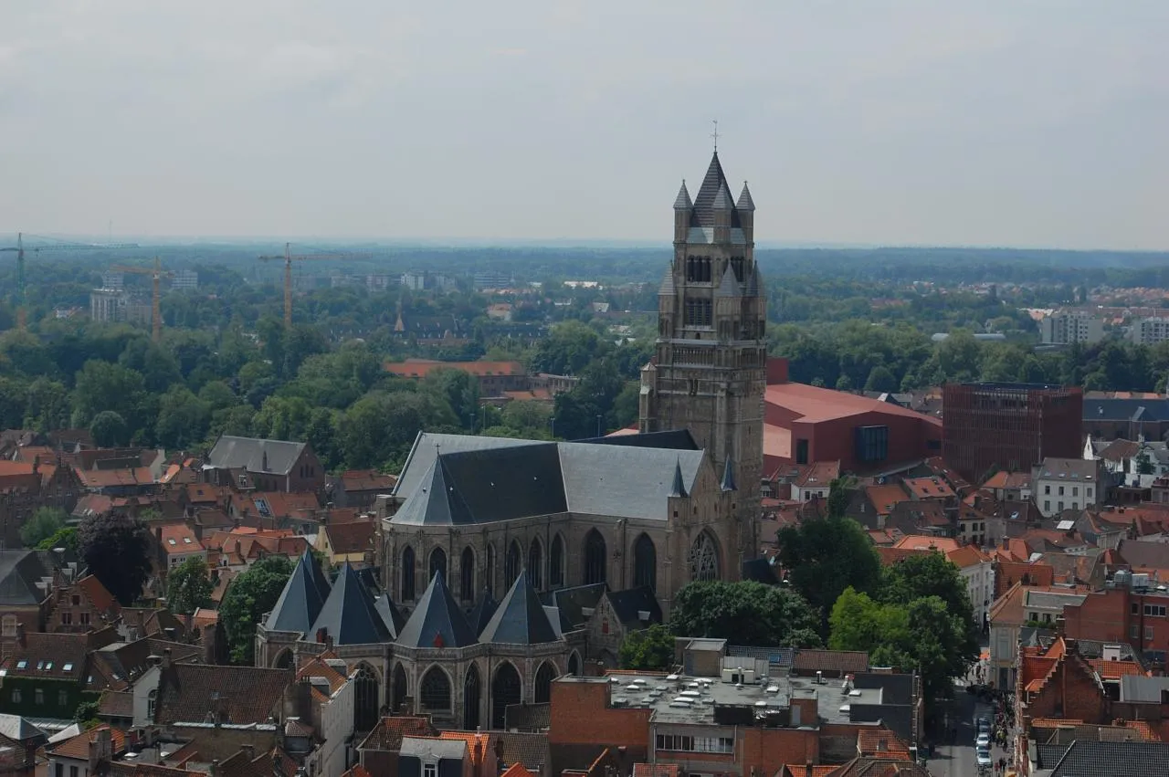 Saint Saviour's Cathedral in Bruges seen from above, its long slate roof and squat brick tower with turreted spires standing among the red-tiled roofs of the surrounding streets