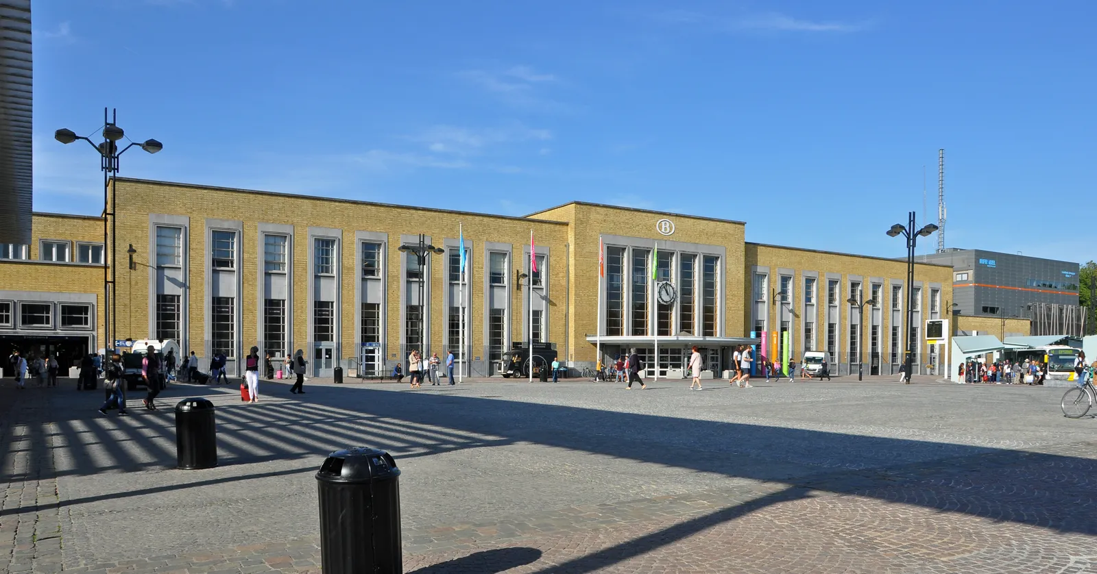 The yellow-brick front of Brugge railway station seen across the open square in front of it, a long symmetrical facade with a raised central block and flagpoles along the roofline