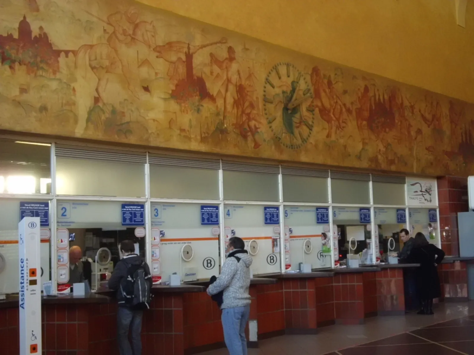 The ticket hall of Brugge railway station, a long painted mural of landscapes and figures running above the row of numbered ticket counters
