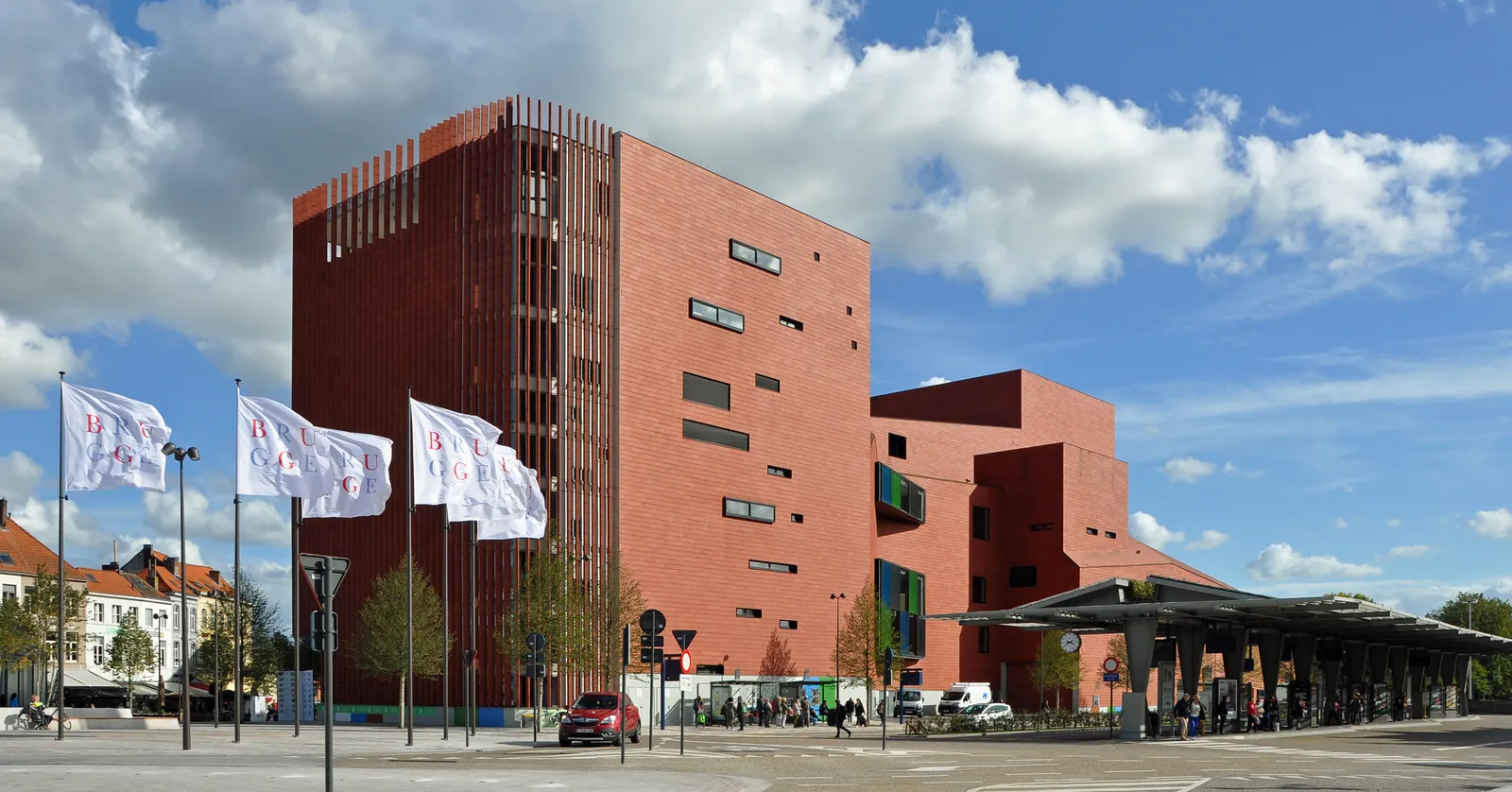 The Bruges concert hall on 't Zand, a tall block clad entirely in dark red terracotta tiles with narrow slot windows, flagpoles and a bus shelter in front of it
