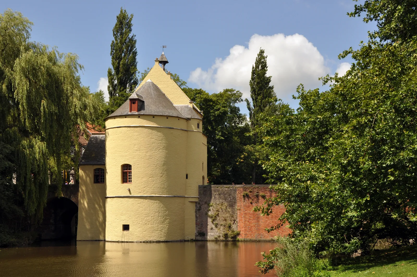The Smedenpoort in Bruges, a pale yellow medieval gatehouse with two round towers and a slate roof standing over the water of the old moat, willows on the bank beside it