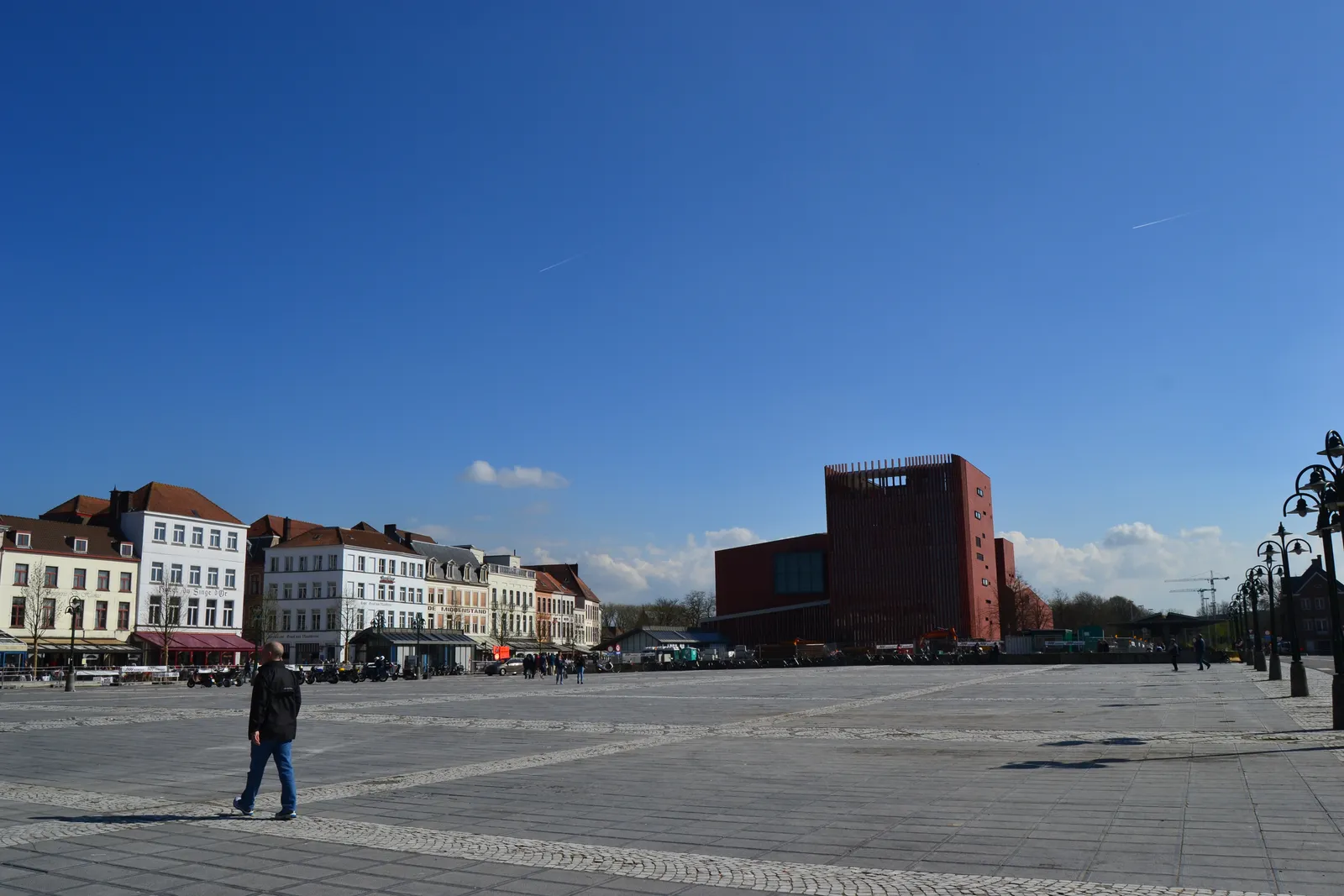 The wide paved expanse of 't Zand in Bruges on a clear day, a row of gabled houses and cafe awnings along the left side and the red terracotta concert hall standing across the far end