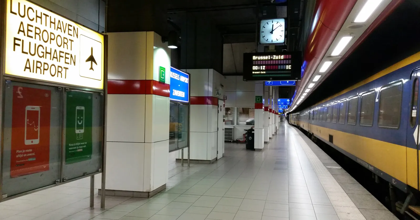 The platform at Brussels Airport's railway station, with an illuminated sign reading Airport in four languages and a blue and yellow train waiting at the platform