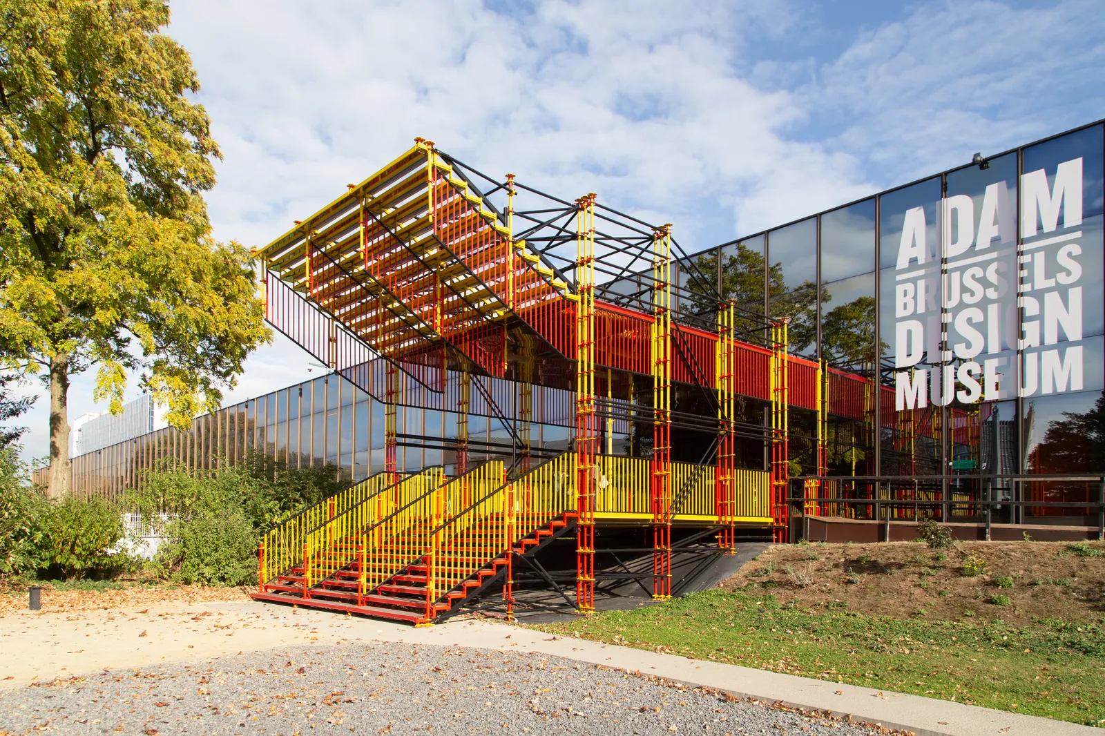 The Design Museum Brussels on the Heysel plateau, a dark glass building with its name in white capitals, fronted by a red and yellow scaffolding staircase installation