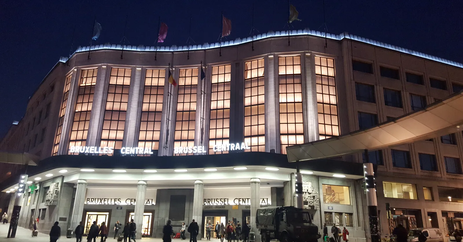 Brussels-Central station lit up after dark, its tall grid of windows glowing above a long canopy carrying the station's name in French and Dutch, with people crossing the square in front