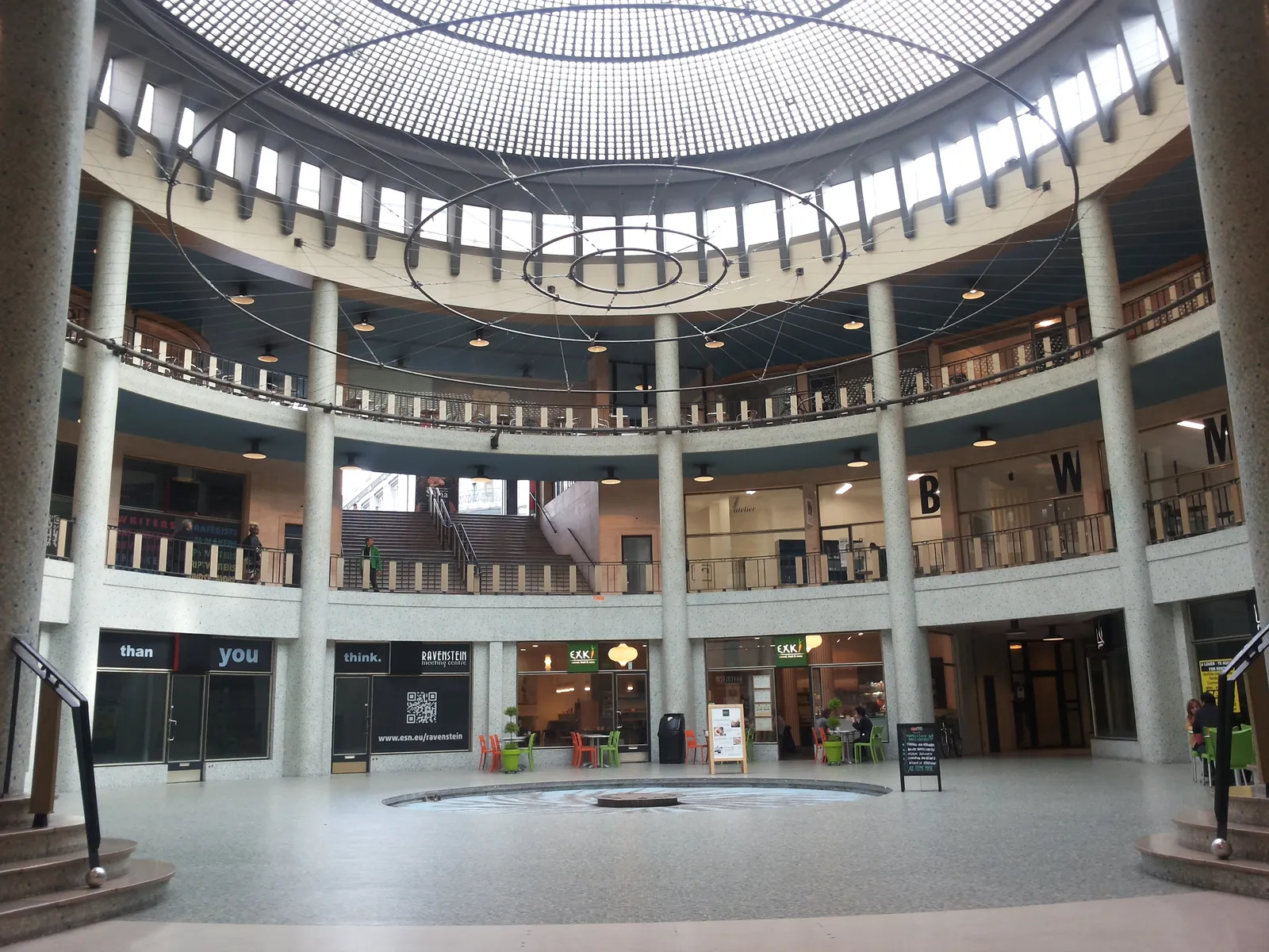 The circular hall of the Galerie Ravenstein in Brussels, a balconied drum of concrete and stone under a wide latticed glass dome, with shopfronts around the ground floor