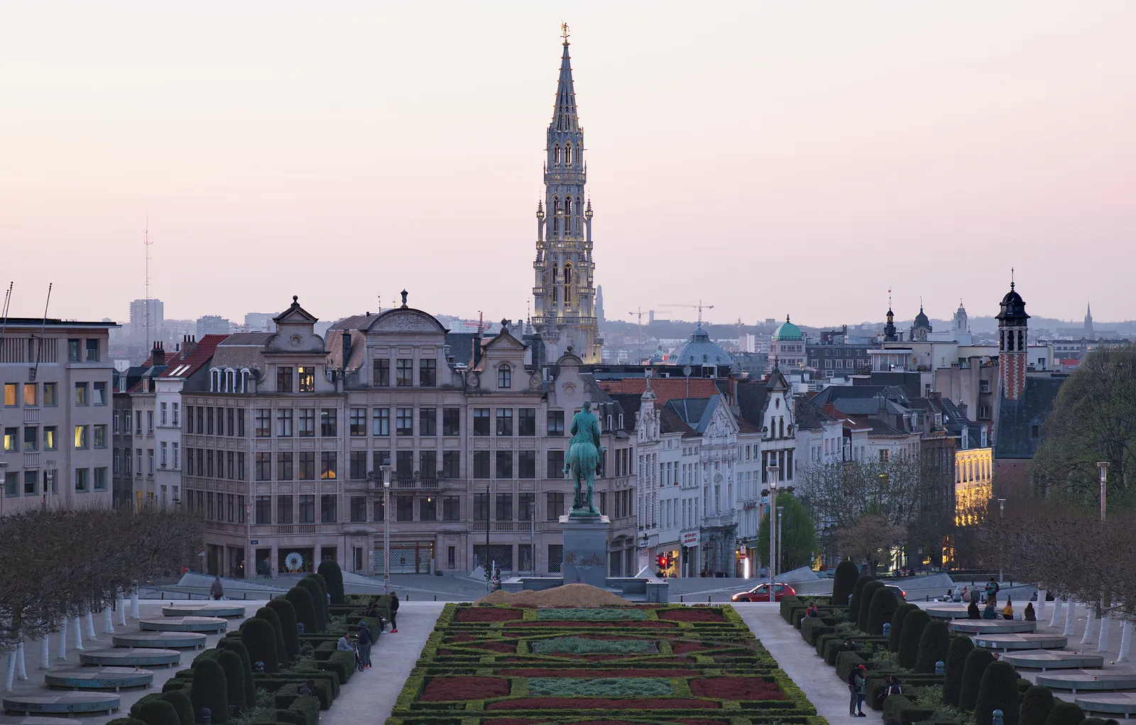 The view from the Mont des Arts terrace in Brussels at dusk, clipped hedges and flowerbeds in the foreground and the Town Hall spire standing above the lower town under a pink sky