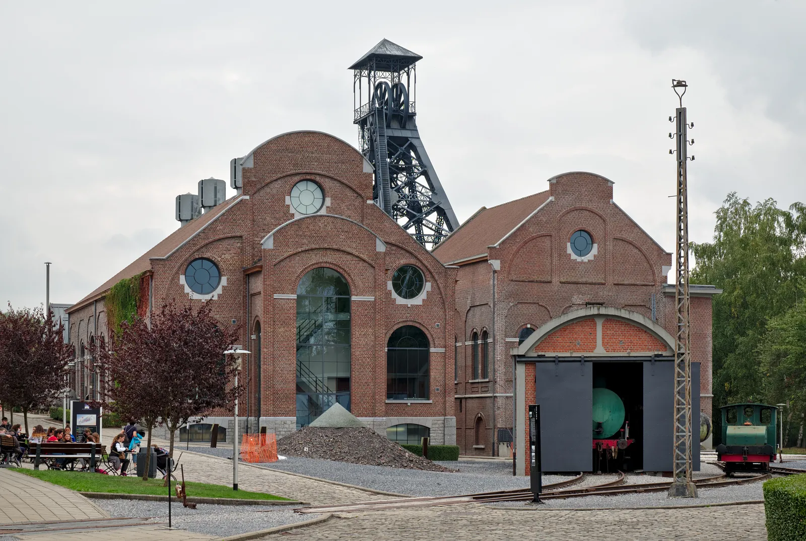 The Bois du Cazier colliery at Marcinelle, brick pithead buildings with round windows under a steel winding tower, with a mine wagon and rails in the yard in front
