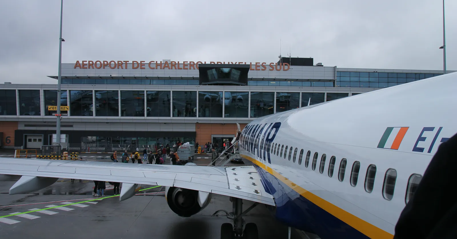 The terminal at Brussels South Charleroi Airport with its name in orange letters across the front, seen past the wing of a parked aircraft on a grey apron