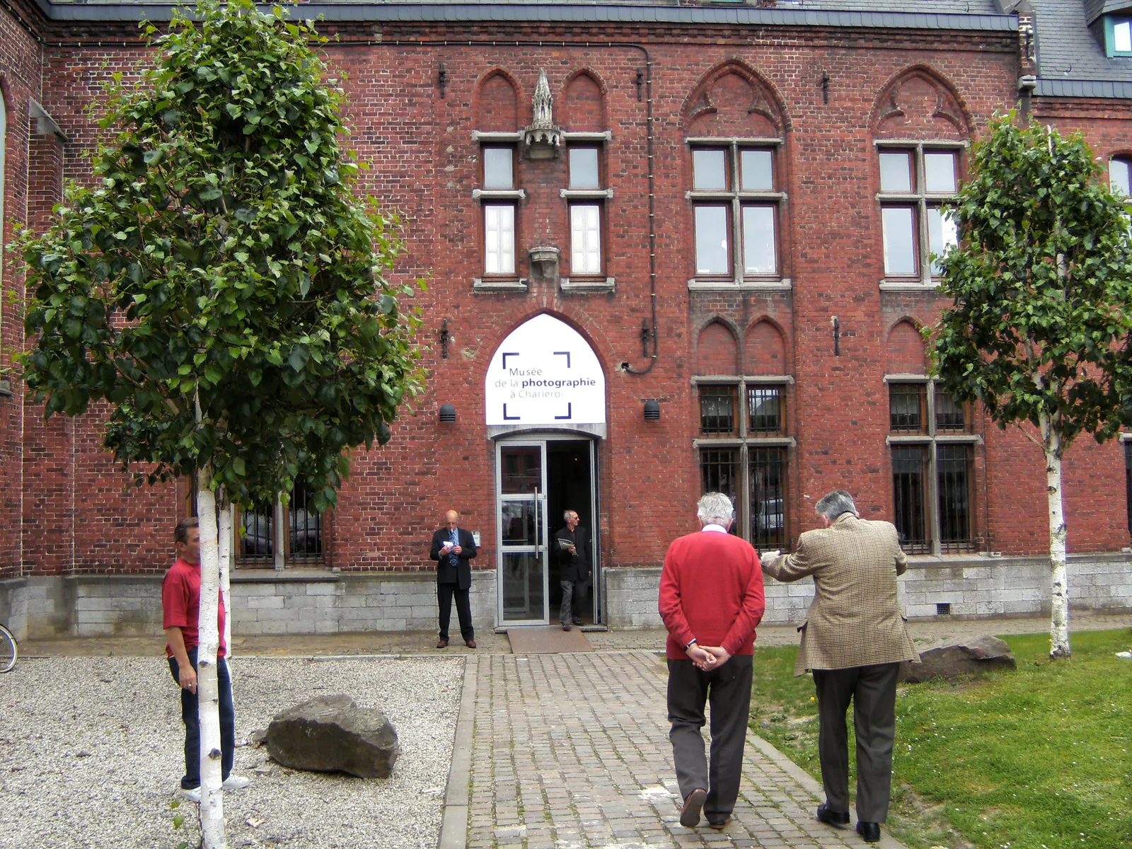 The entrance to the Museum of Photography at Mont-sur-Marchienne, a red brick neo-Gothic convent building with pointed windows and a glass porch, with visitors walking up the path
