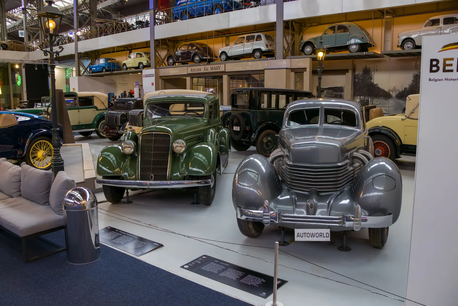Vintage cars lined up on the floor of Autoworld in Brussels, a 1930s saloon and a streamlined silver coupe in the foreground beneath a mezzanine of more cars and iron roof trusses