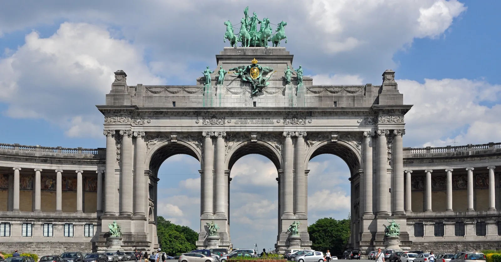 The triumphal arch in the Parc du Cinquantenaire in Brussels, three stone arches carrying a green bronze chariot group, with colonnaded wings curving away on both sides under a blue sky