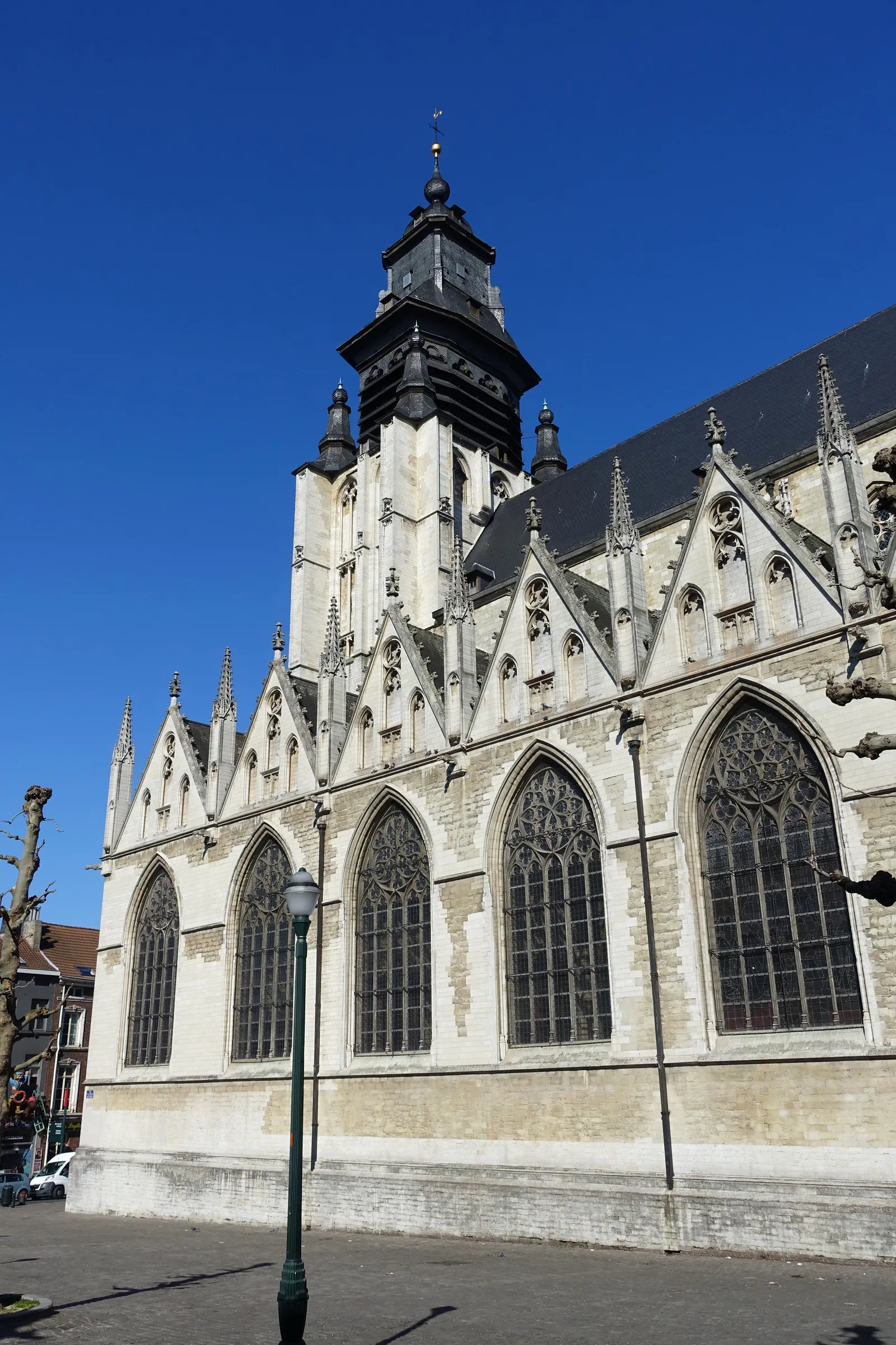 The pale stone flank of Notre-Dame de la Chapelle in Brussels, a row of Gothic windows and gabled buttresses under a black baroque bell tower against a clear blue sky