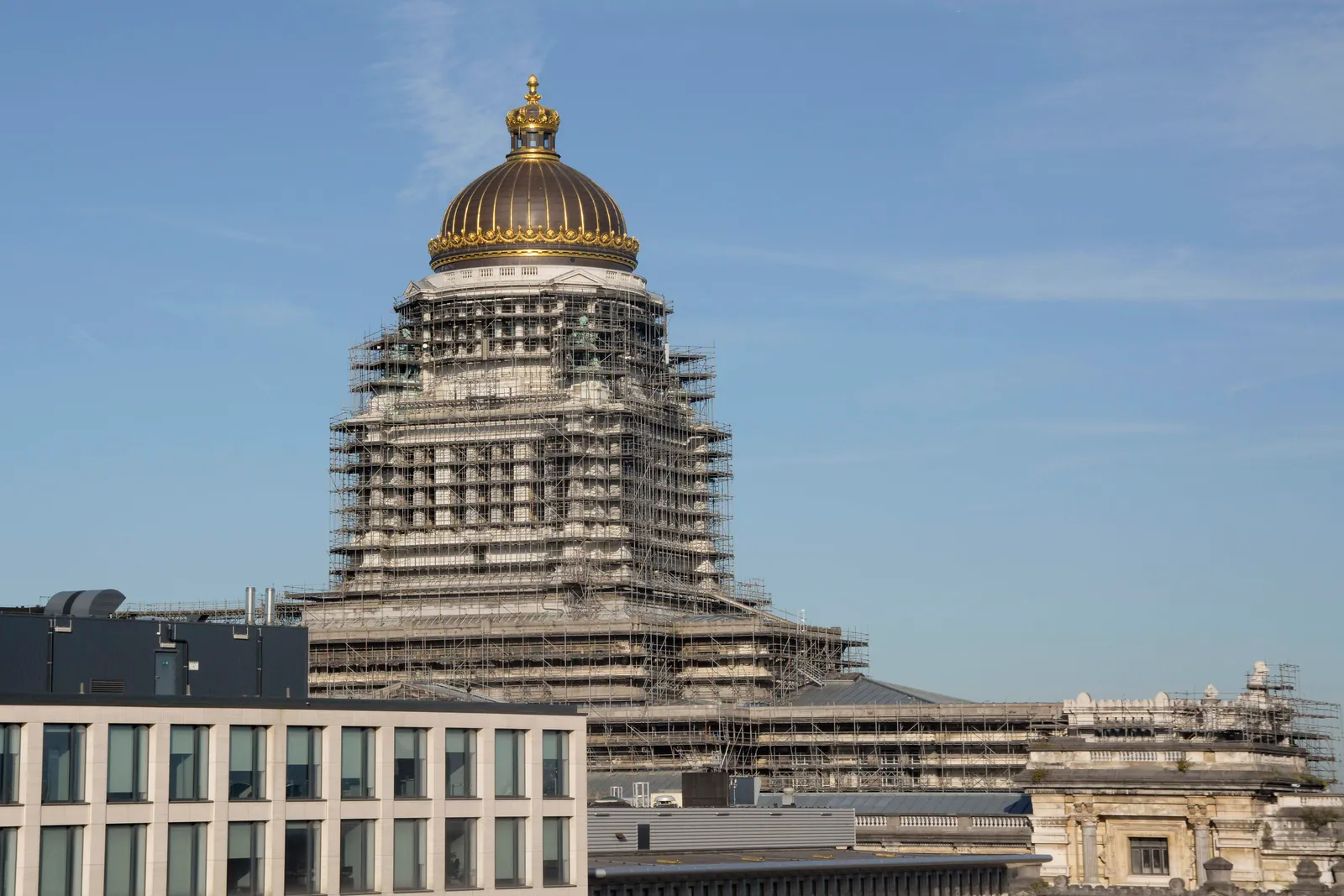 The dome of the Brussels Palace of Justice wrapped in tiers of steel scaffolding, its gilded crown still showing at the top, seen over the rooftops against a blue sky