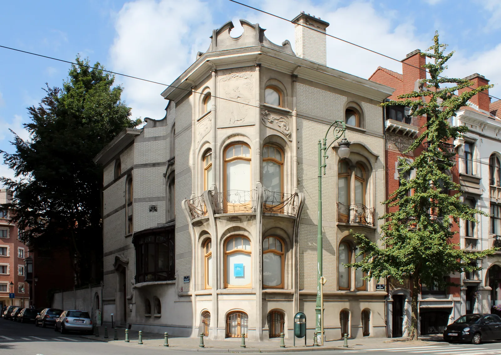 The Hannon house in Saint-Gilles, a corner townhouse with curved Art Nouveau window bays, a wrought-iron balcony and pale stonework, standing at a junction of two tree-lined avenues