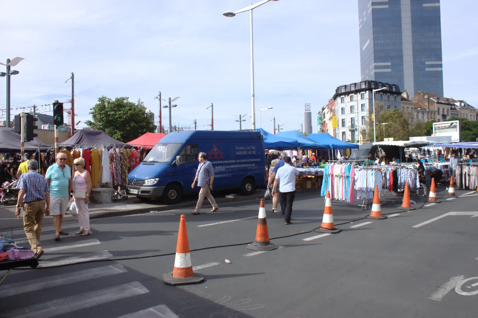 Market stalls and clothing rails set up along a wide boulevard beside Brussels-Midi station, with delivery vans parked between the awnings and shoppers crossing the road