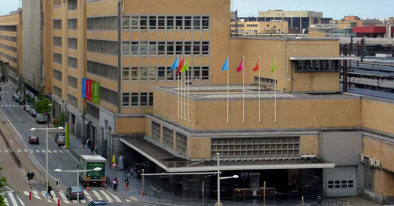 The yellow brick bulk of Brussels-Midi station seen from above, flags along its roofline, a metro entrance beneath it and tram tracks curving along the boulevard in front